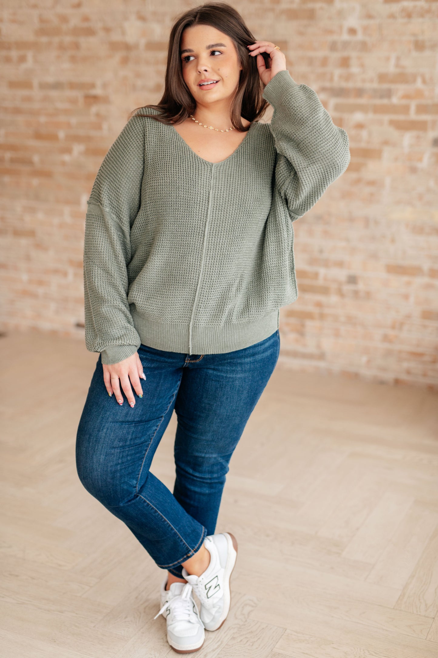 Woman wearing a green sweater and blue jeans with white sneakers against a brick wall.