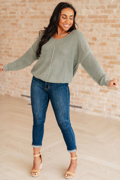 Woman wearing a green sweater and blue jeans standing against a brick wall.