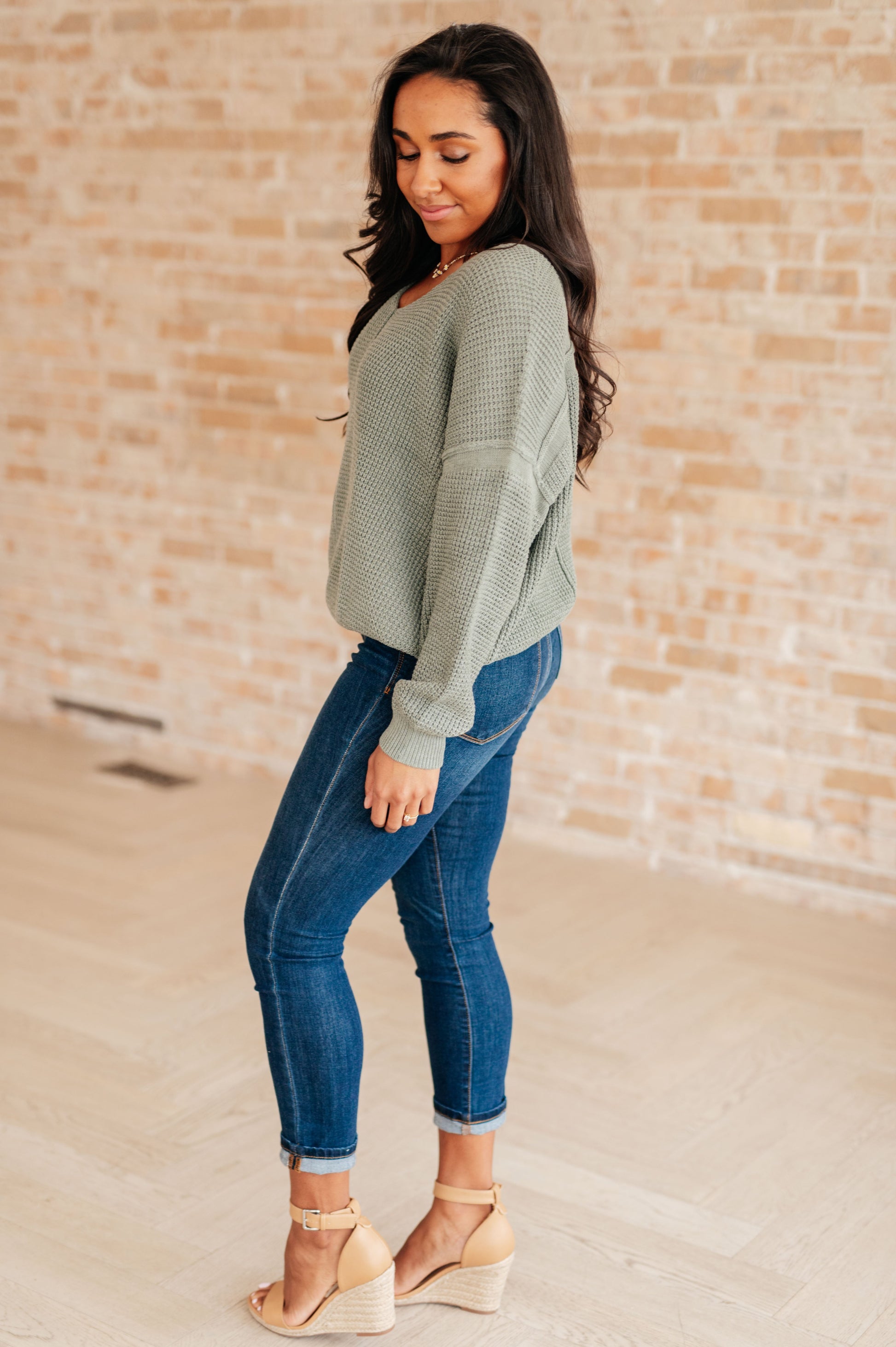 Woman wearing a green sweater and blue jeans standing against a brick wall.