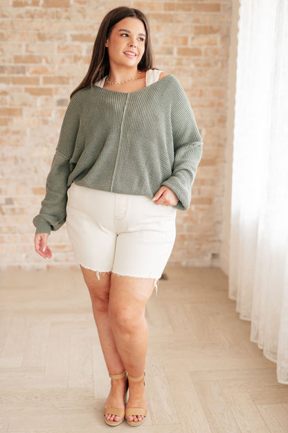 Woman wearing a green sweater and white shorts standing in a room with a brick wall and white curtains.