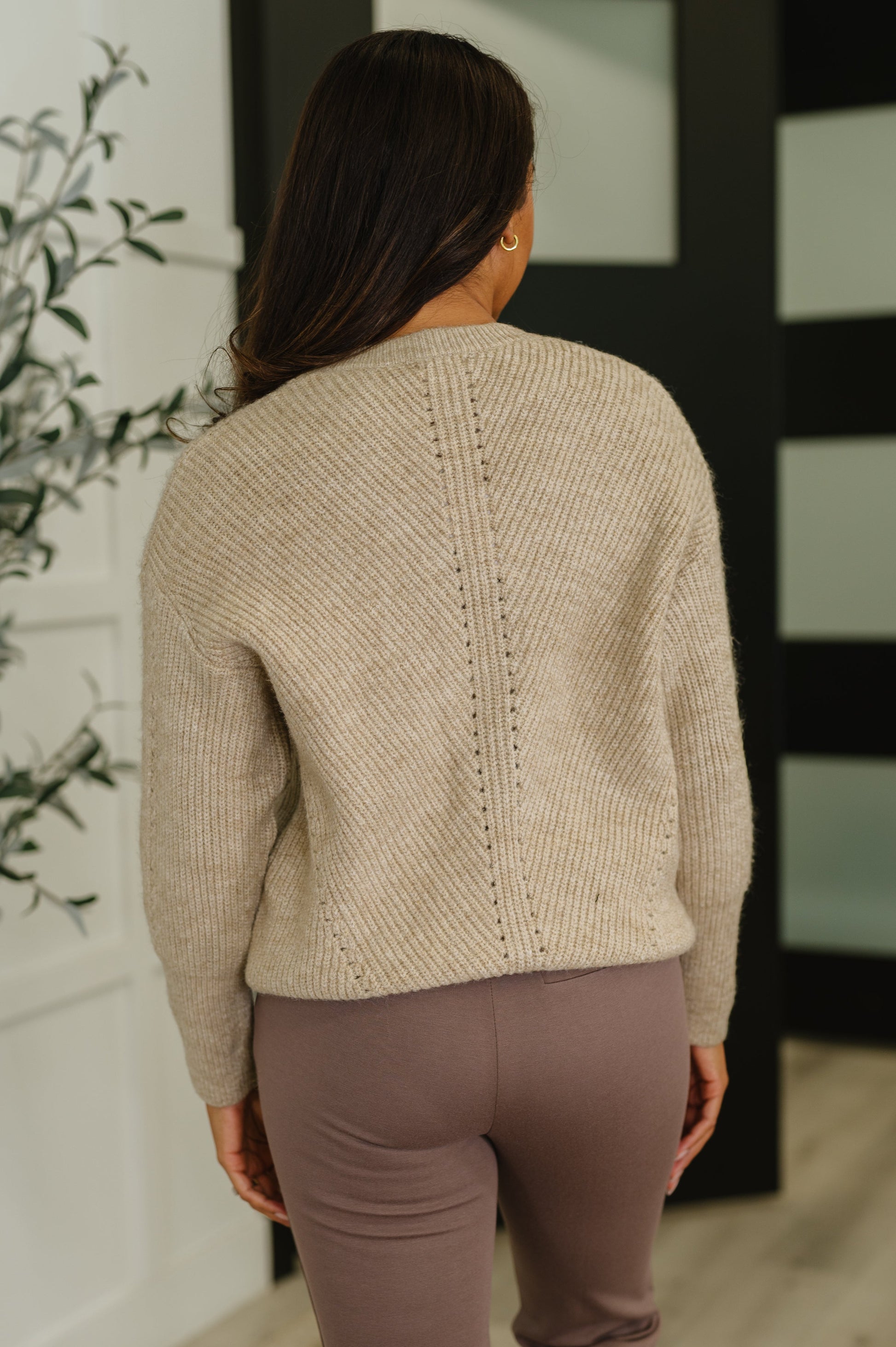 Person wearing a beige sweater and brown pants standing in a room with a plant and black screen in the background.
