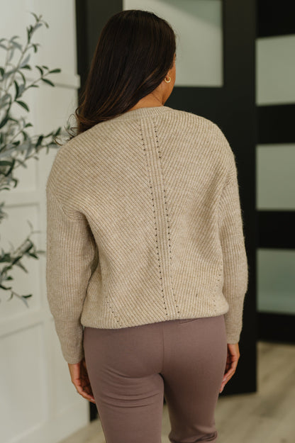 Person wearing a beige sweater and brown pants standing in a room with a plant and black screen in the background.