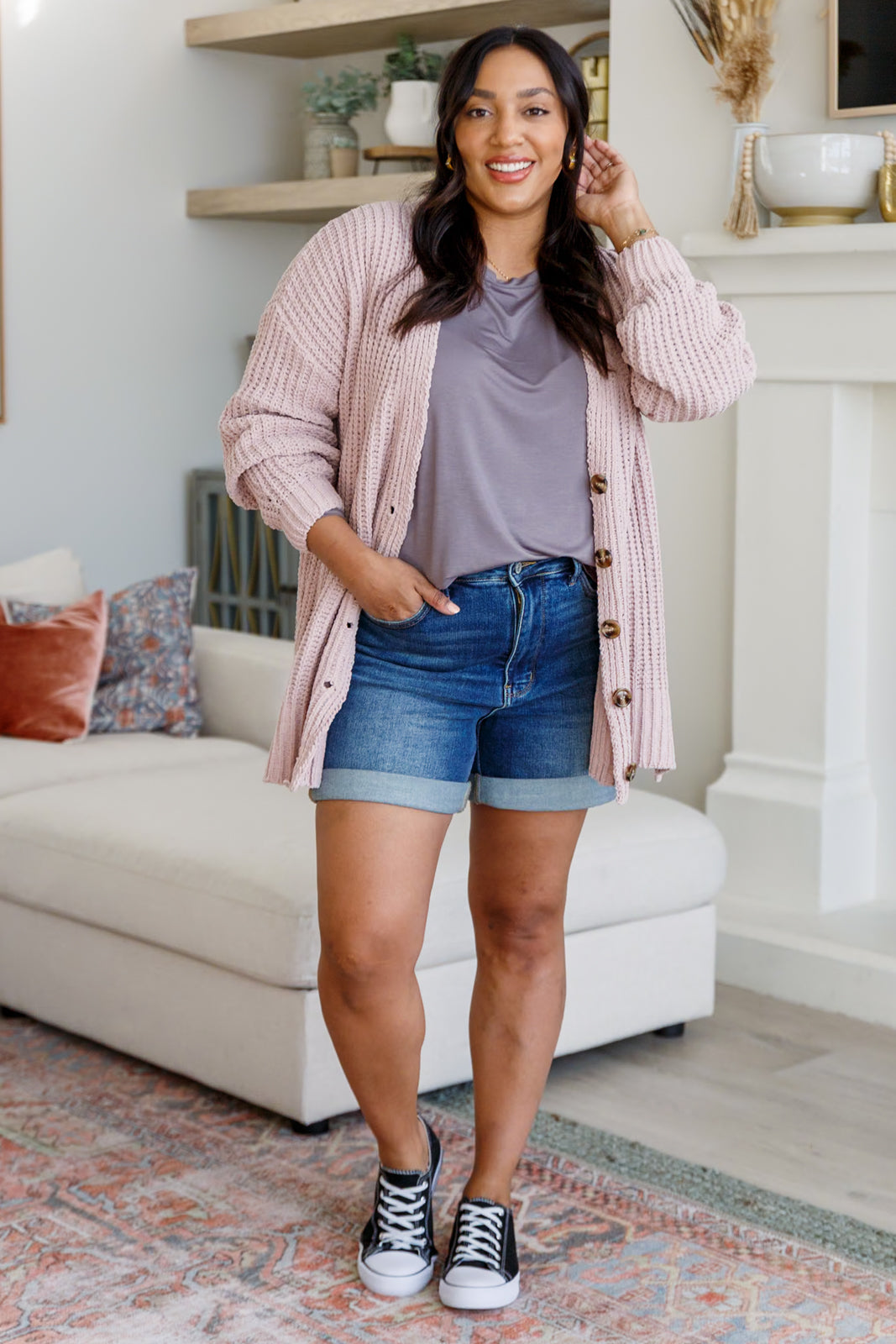 Woman standing in a living room wearing a pink cardigan, gray shirt, denim shorts, and black sneakers.