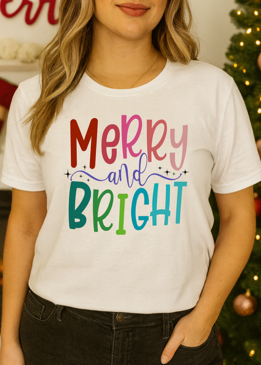 Person wearing a white t-shirt with 'Merry and Bright' text in colorful letters, standing in front of a decorated Christmas tree.