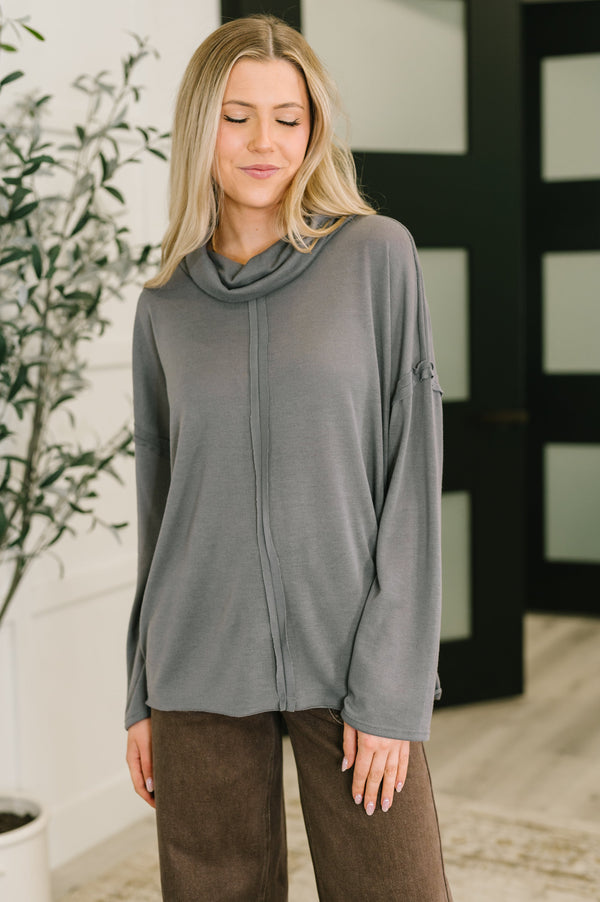 Woman wearing a gray long-sleeve top with a cowl neck in an indoor setting.