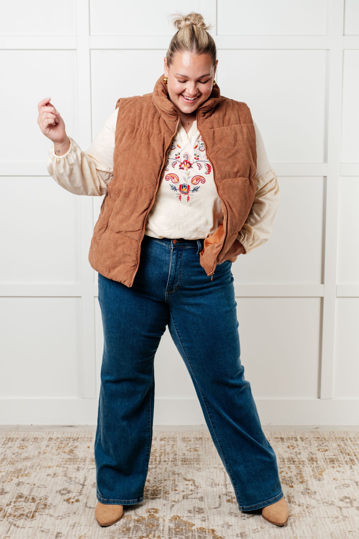 Person wearing a brown vest, white shirt with floral design, and blue jeans against a white wall.