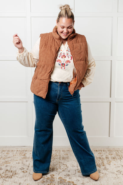 Person wearing a brown vest, white shirt with floral design, and blue jeans against a white wall.