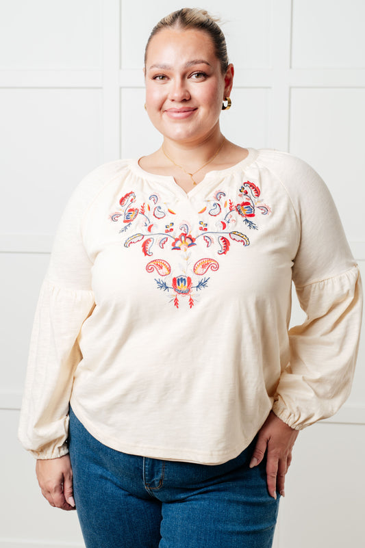 Woman wearing a cream blouse with colorful embroidery against a white tiled wall.