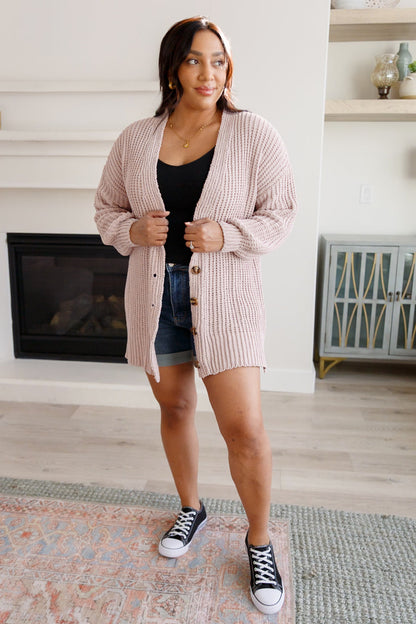 Woman wearing a pink cardigan, black top, denim shorts, and black sneakers in a living room.