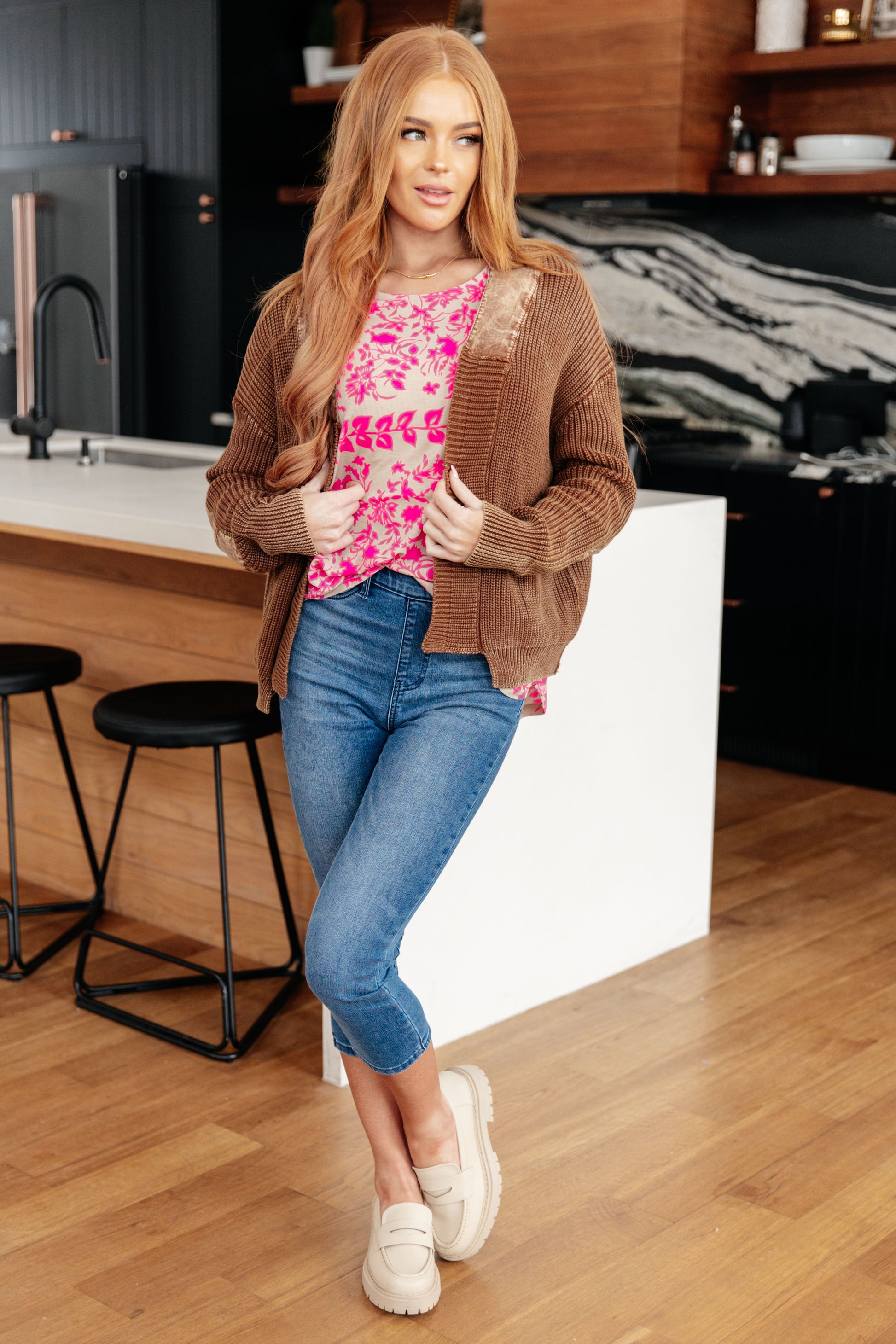 Woman in a kitchen wearing a brown cardigan, pink floral top, blue jeans, and white shoes.