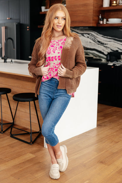 Woman in a kitchen wearing a brown cardigan, pink floral top, blue jeans, and white shoes.