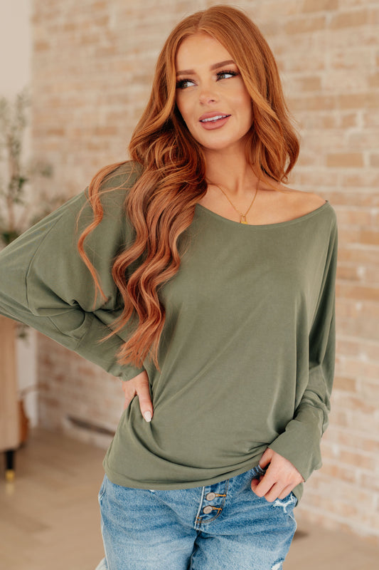 Woman wearing an olive green long-sleeve top and blue jeans against a brick wall.