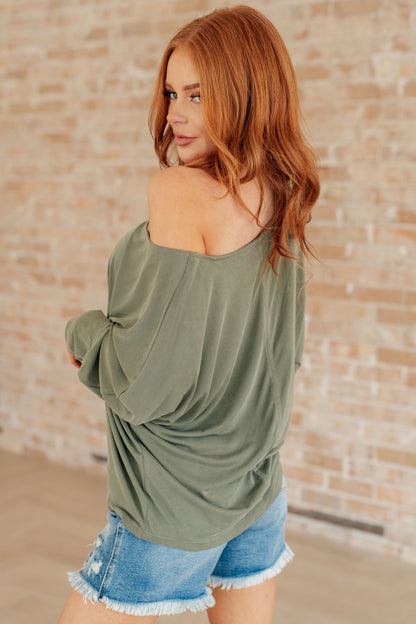 Woman wearing an olive green off-shoulder top and denim shorts against a brick wall.
