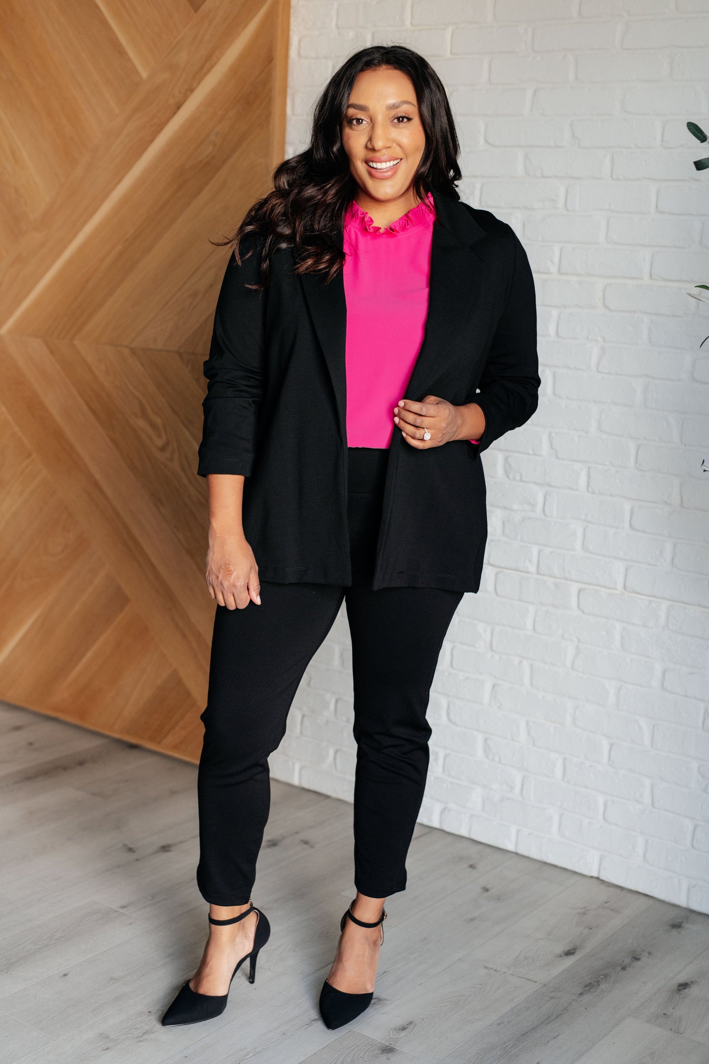 Woman in a black blazer and pink shirt standing against a white brick wall.