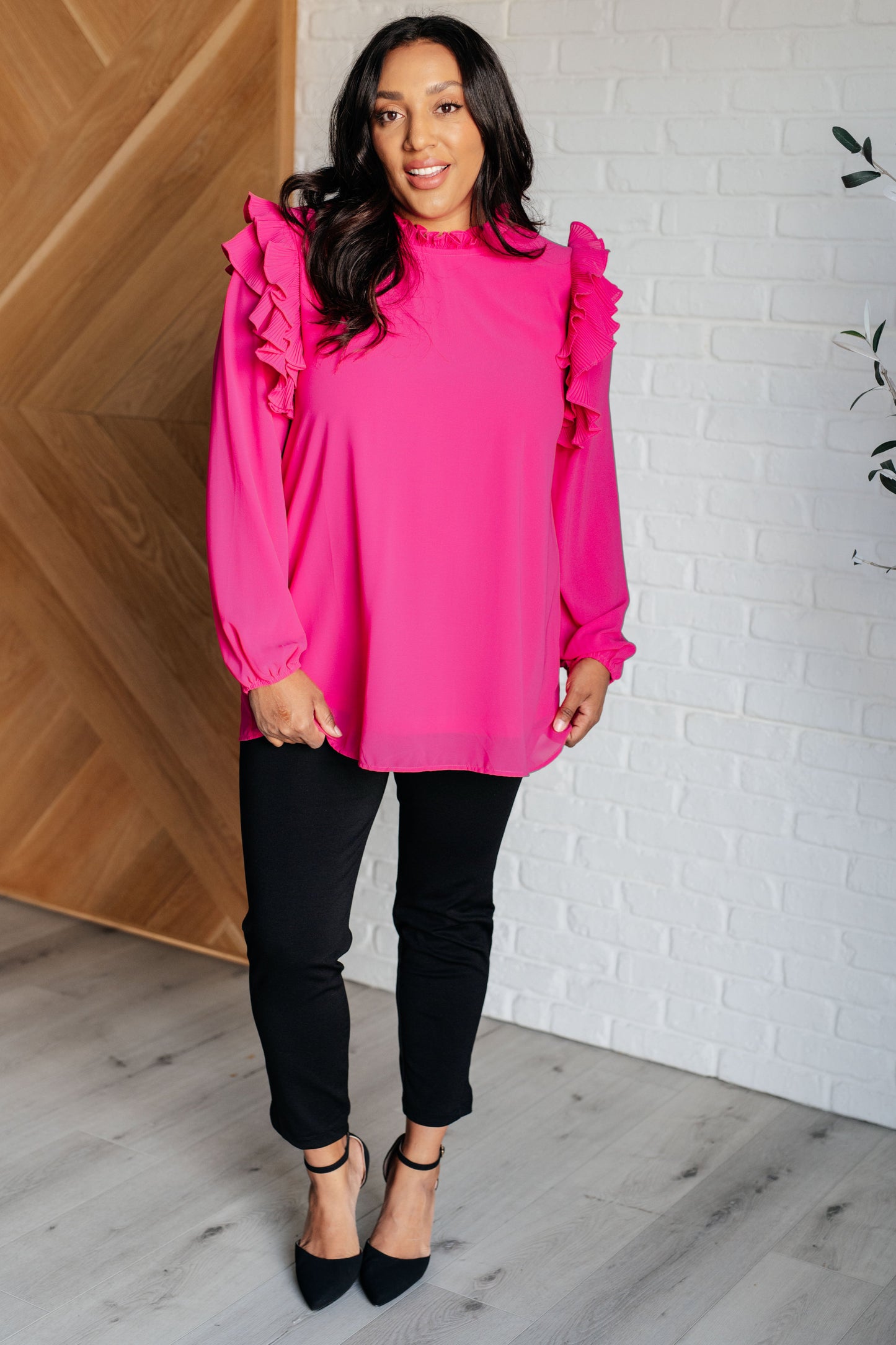 Woman wearing a bright pink blouse with ruffled sleeves and black pants indoors.