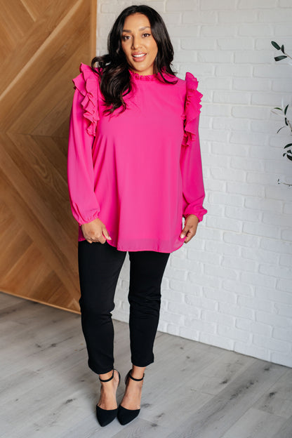 Woman wearing a bright pink blouse with ruffled sleeves and black pants indoors.