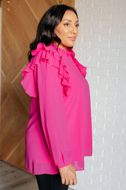 Woman wearing a bright pink blouse with ruffled sleeves against a white brick wall.