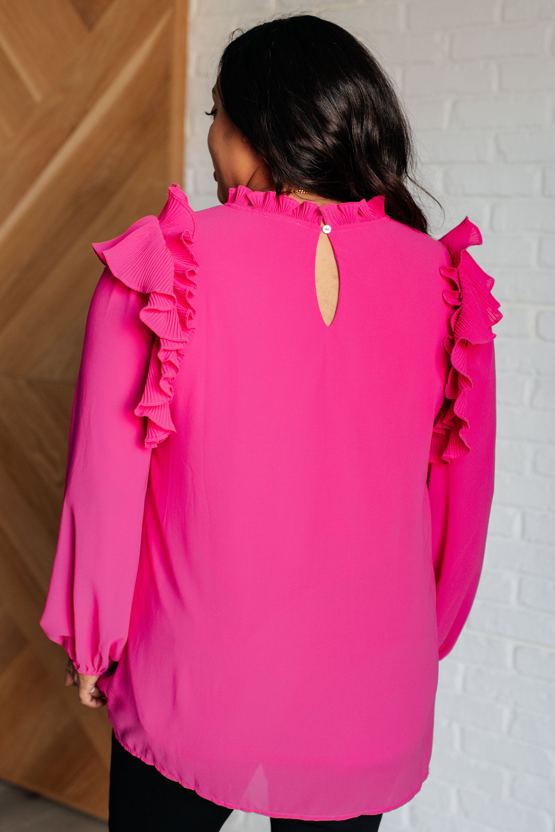 Woman wearing a bright pink blouse with ruffled sleeves against a white brick wall.