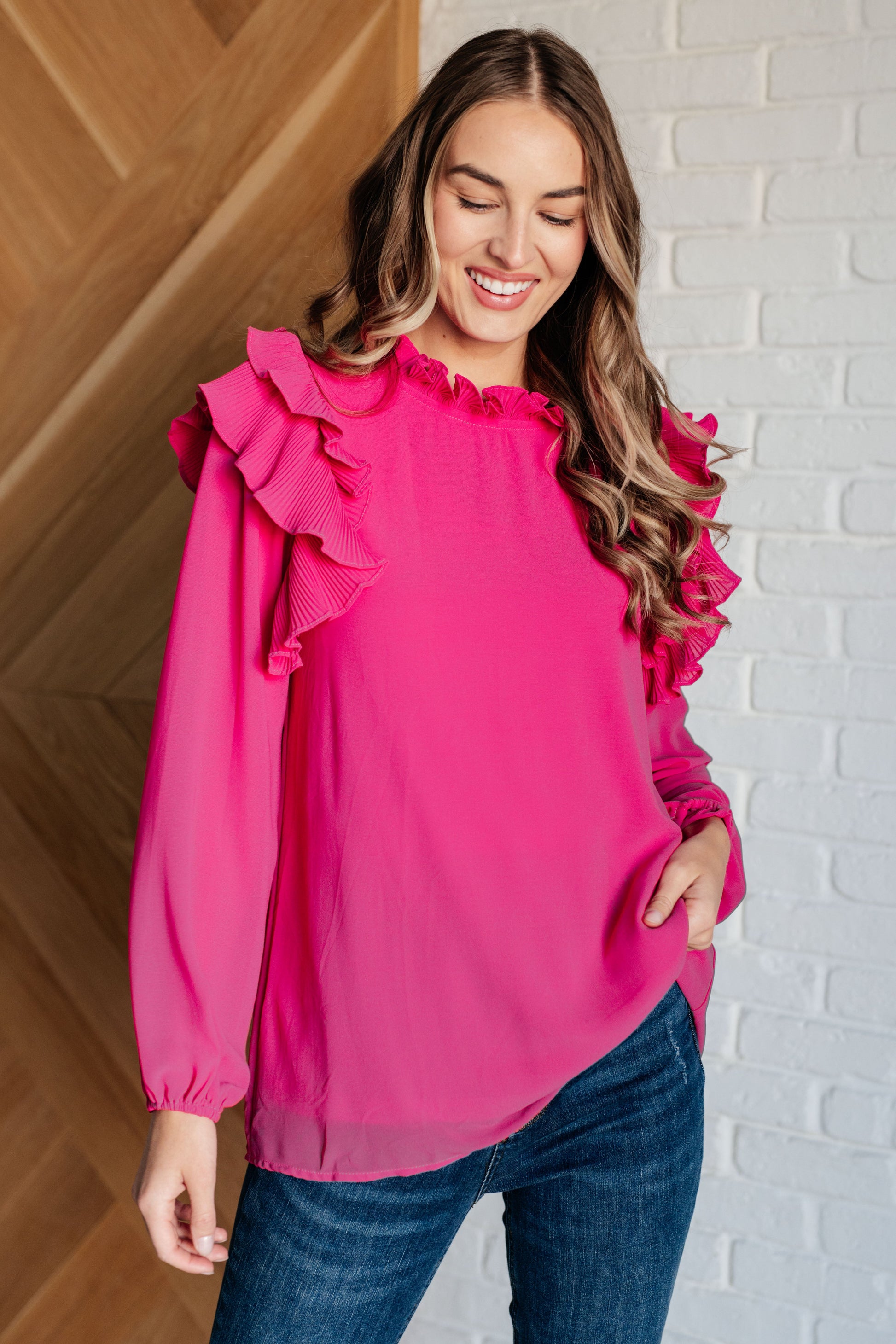 Woman wearing a pink blouse with ruffled sleeves against a white brick wall.