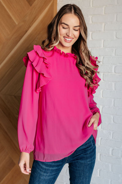 Woman wearing a pink blouse with ruffled sleeves against a white brick wall.