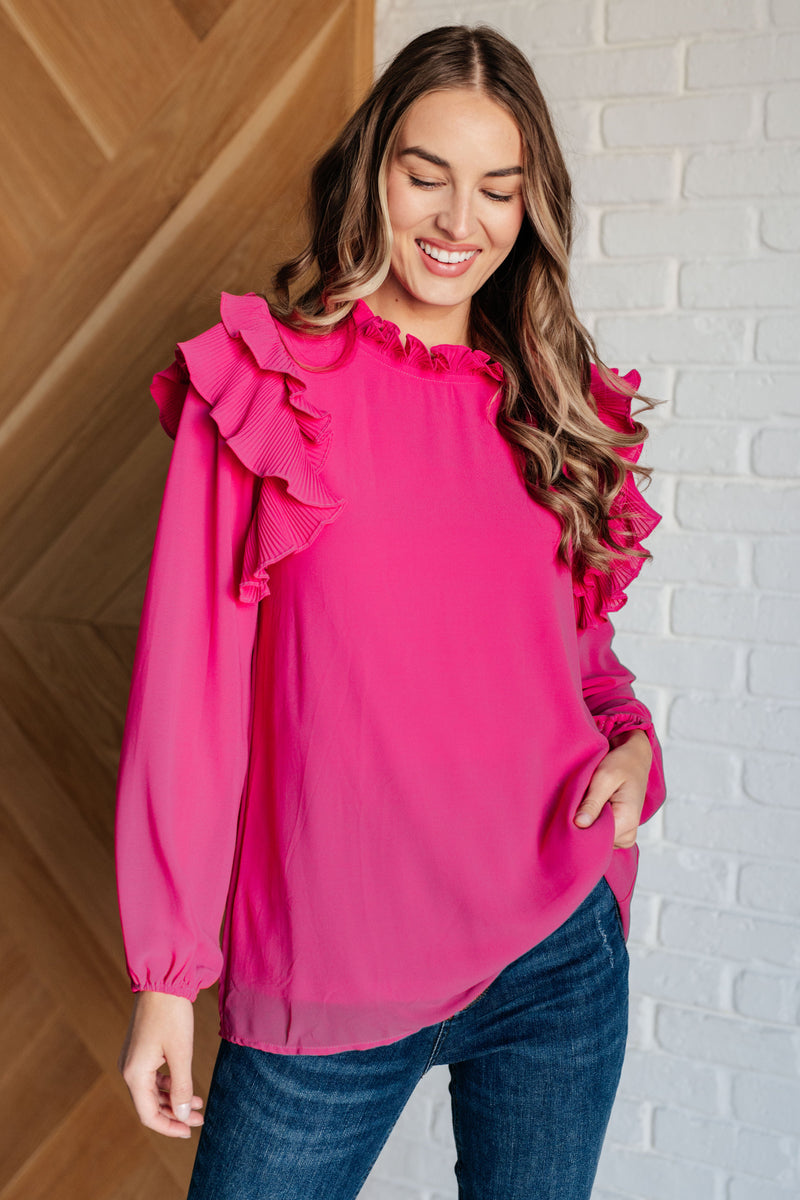 Woman wearing a pink blouse with ruffled sleeves against a white brick wall.