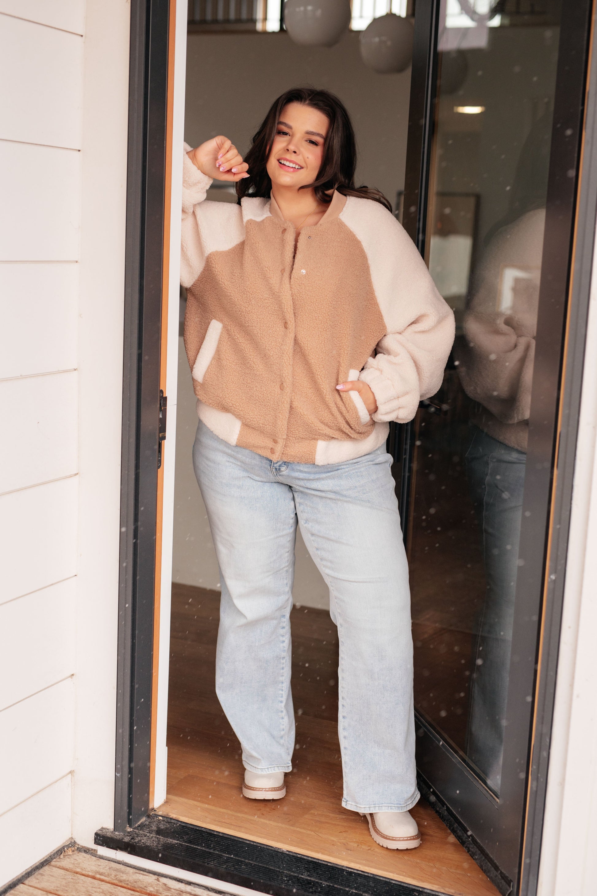 Woman standing in a doorway wearing a beige and white jacket and light blue jeans.