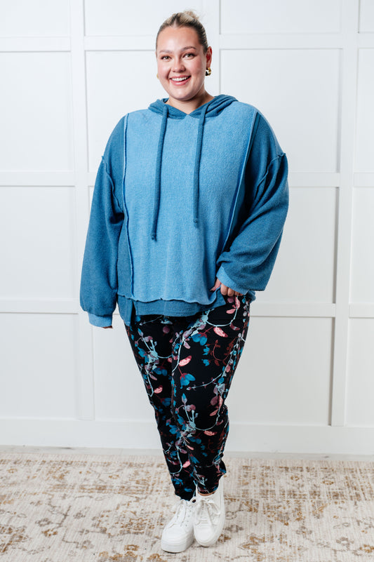 Person wearing a blue hoodie and patterned pants standing against a white wall.