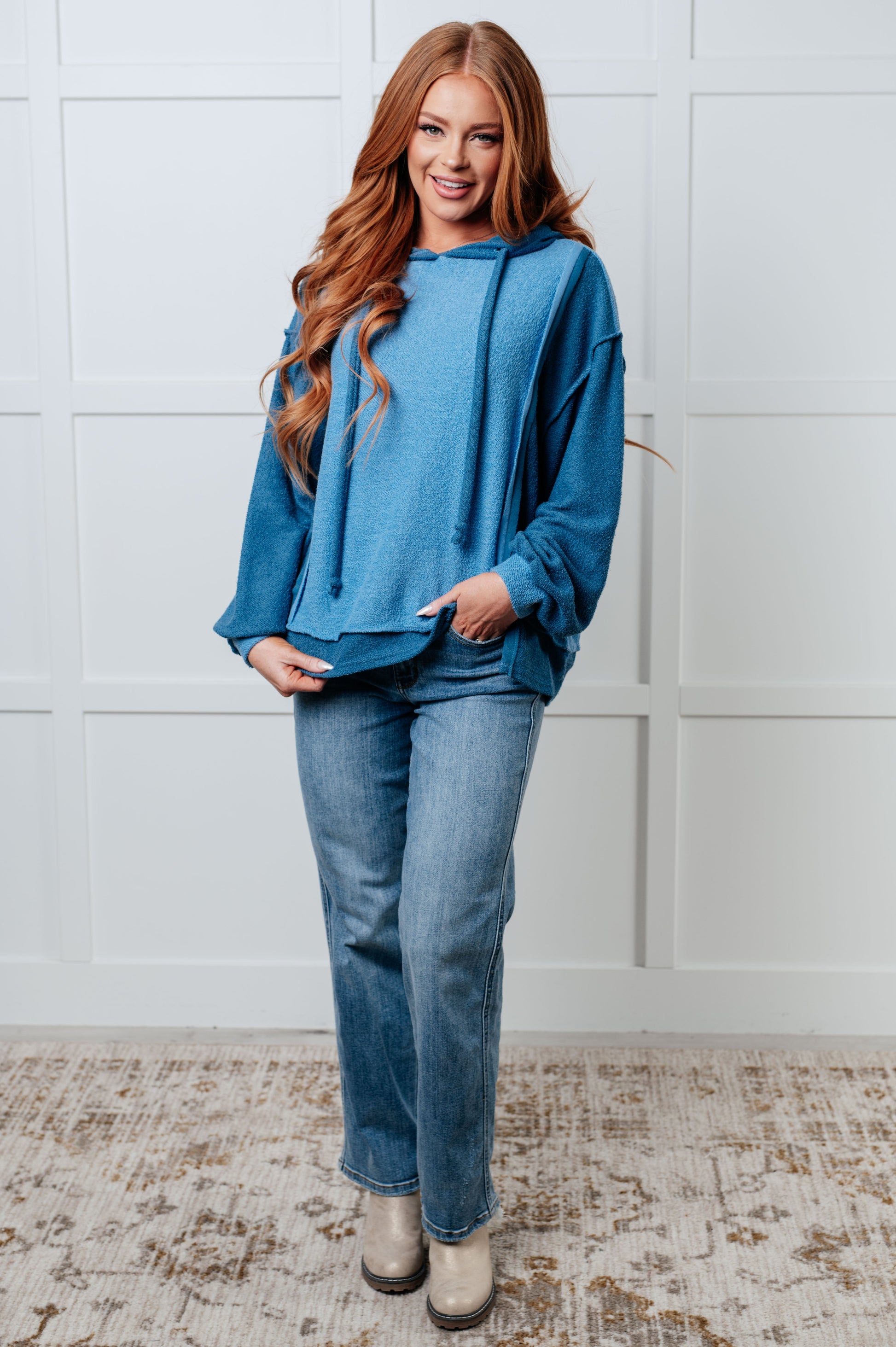 Woman wearing a blue hoodie and jeans standing against a white paneled wall.