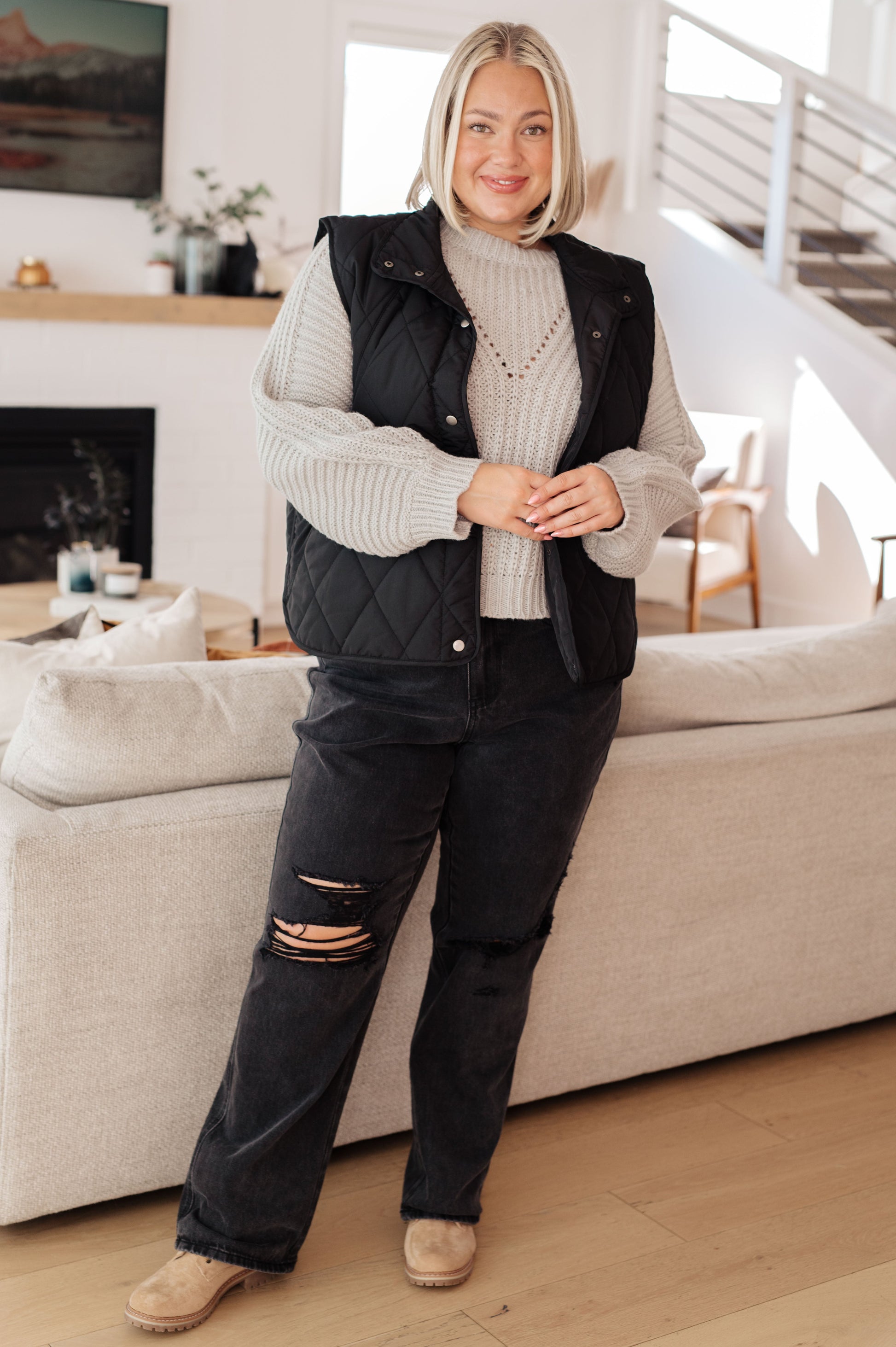 Woman standing in a living room wearing a black vest and ripped jeans.