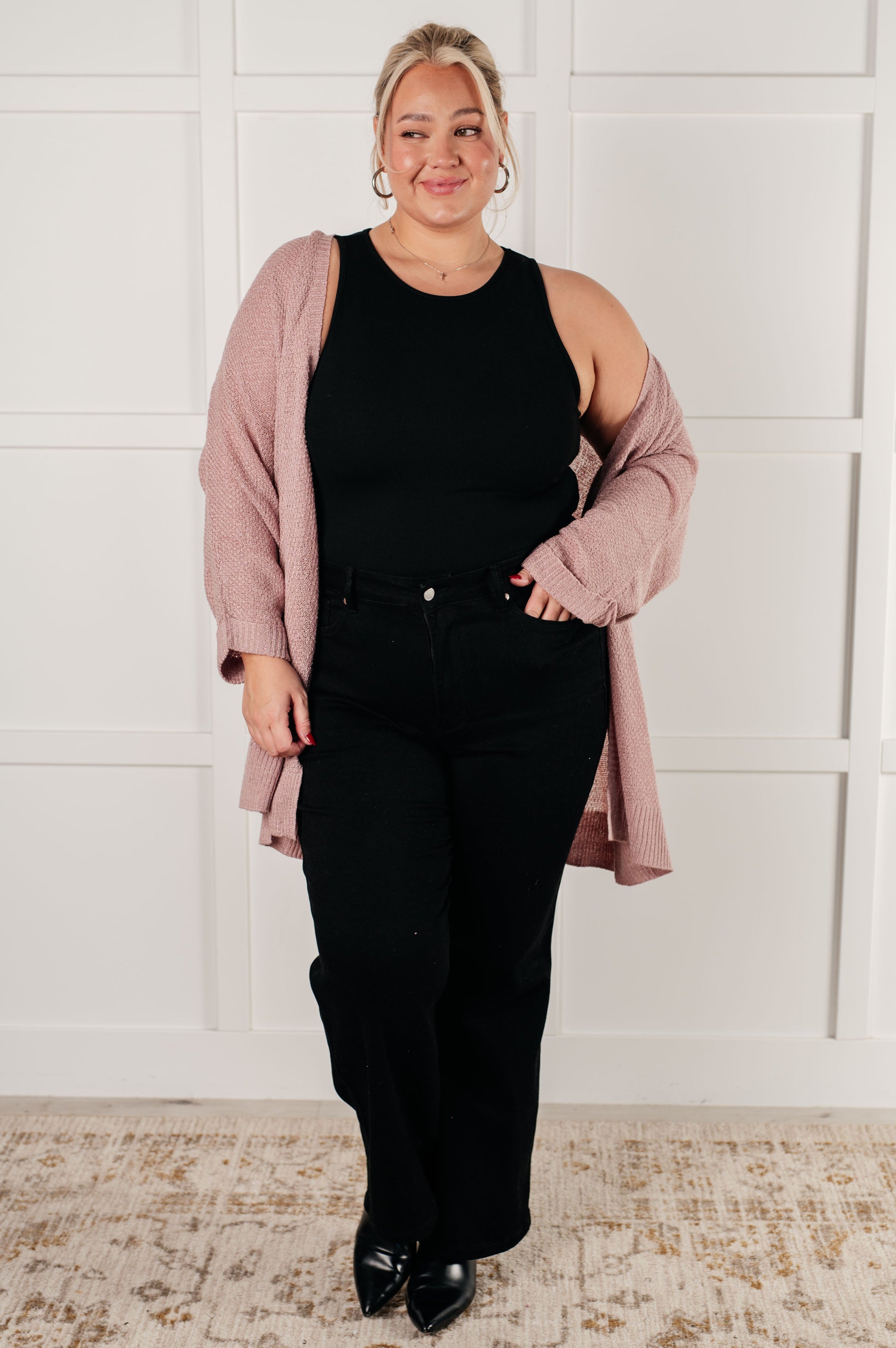 Woman wearing a black sleeveless top and black pants with a pink cardigan against a white paneled wall.