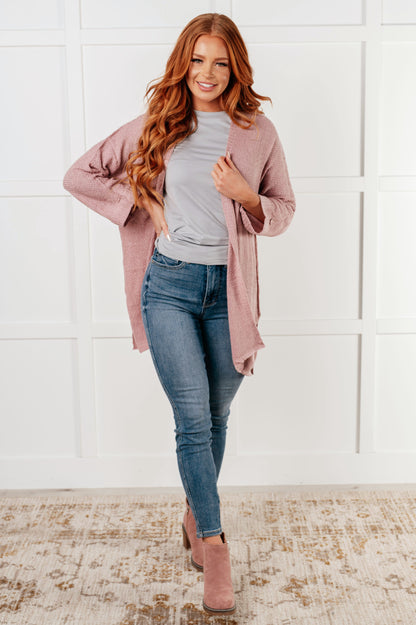 Woman wearing a pink cardigan, gray shirt, and blue jeans against a white wall.