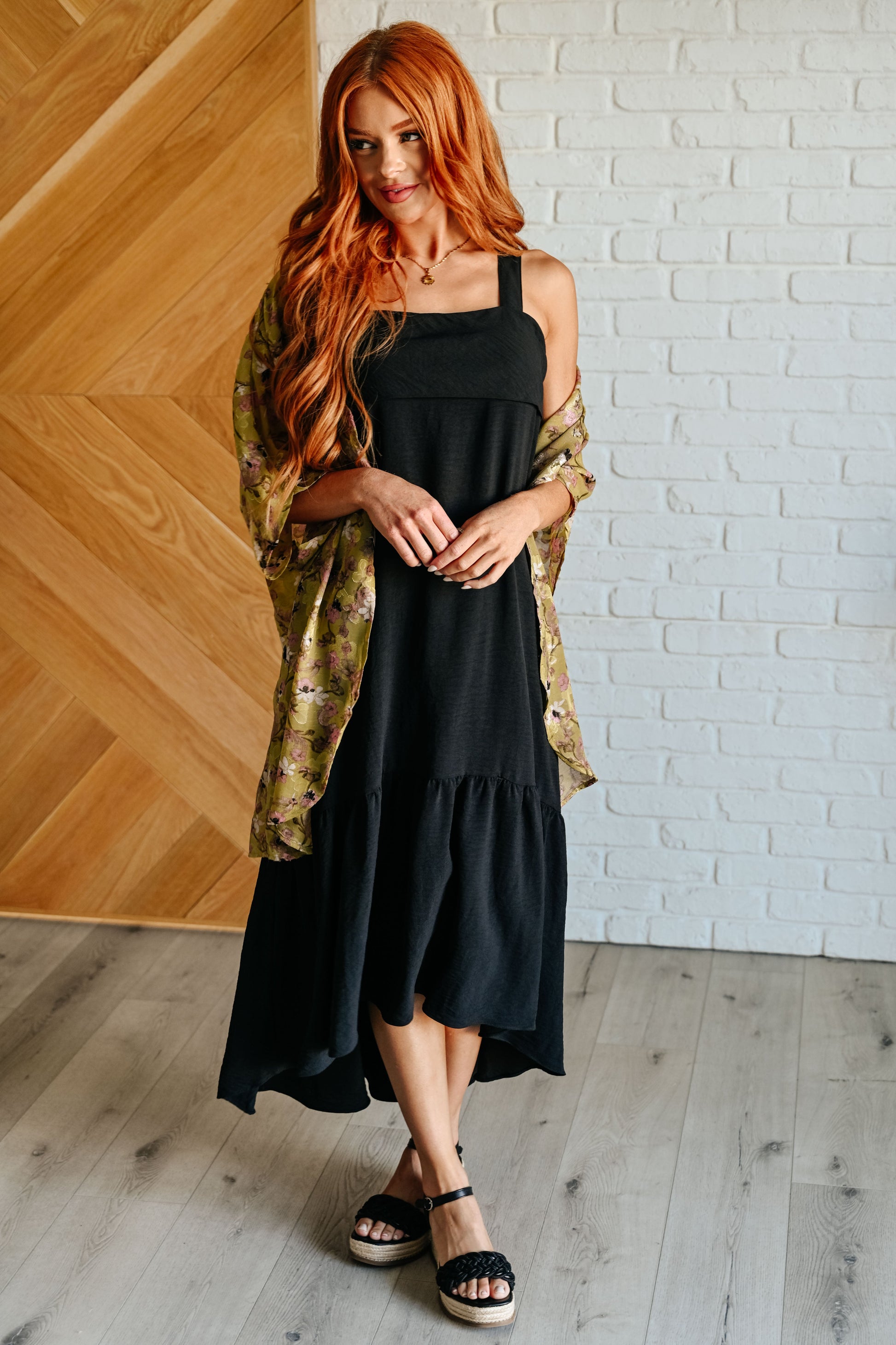 Woman wearing a black dress with a green patterned shawl in a room with wooden and white brick walls.