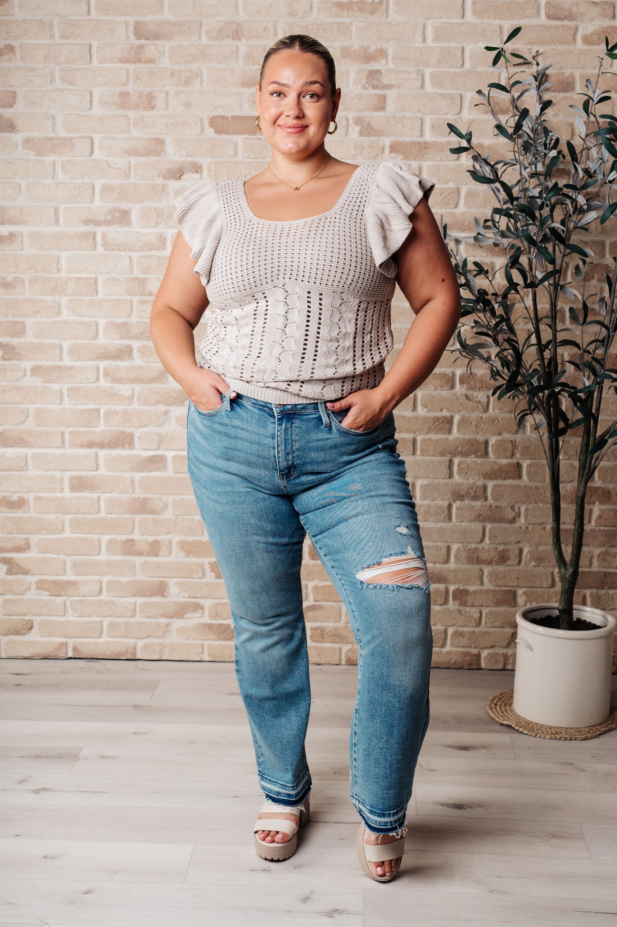 Woman wearing a light gray top and blue jeans standing against a brick wall.