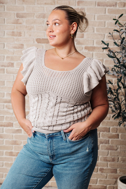 Woman wearing a beige crochet top and blue jeans against a brick wall.