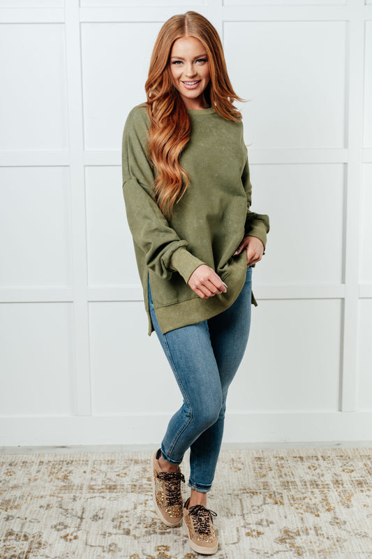 Woman wearing a green sweater and blue jeans standing against a white wall.