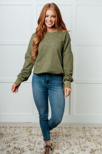 Woman wearing an olive green sweatshirt and blue jeans standing against a white wall.