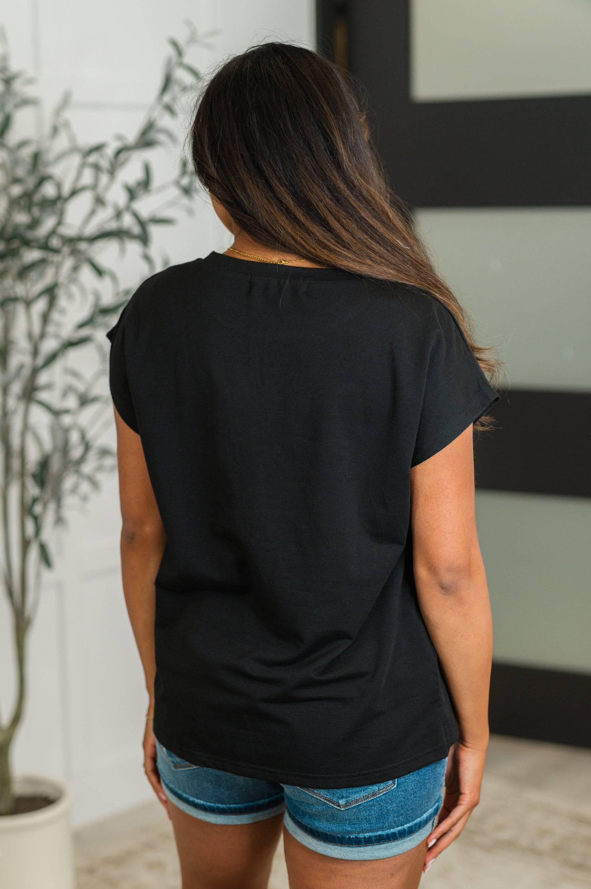 Person wearing a black t-shirt and blue shorts standing indoors with a plant in the background