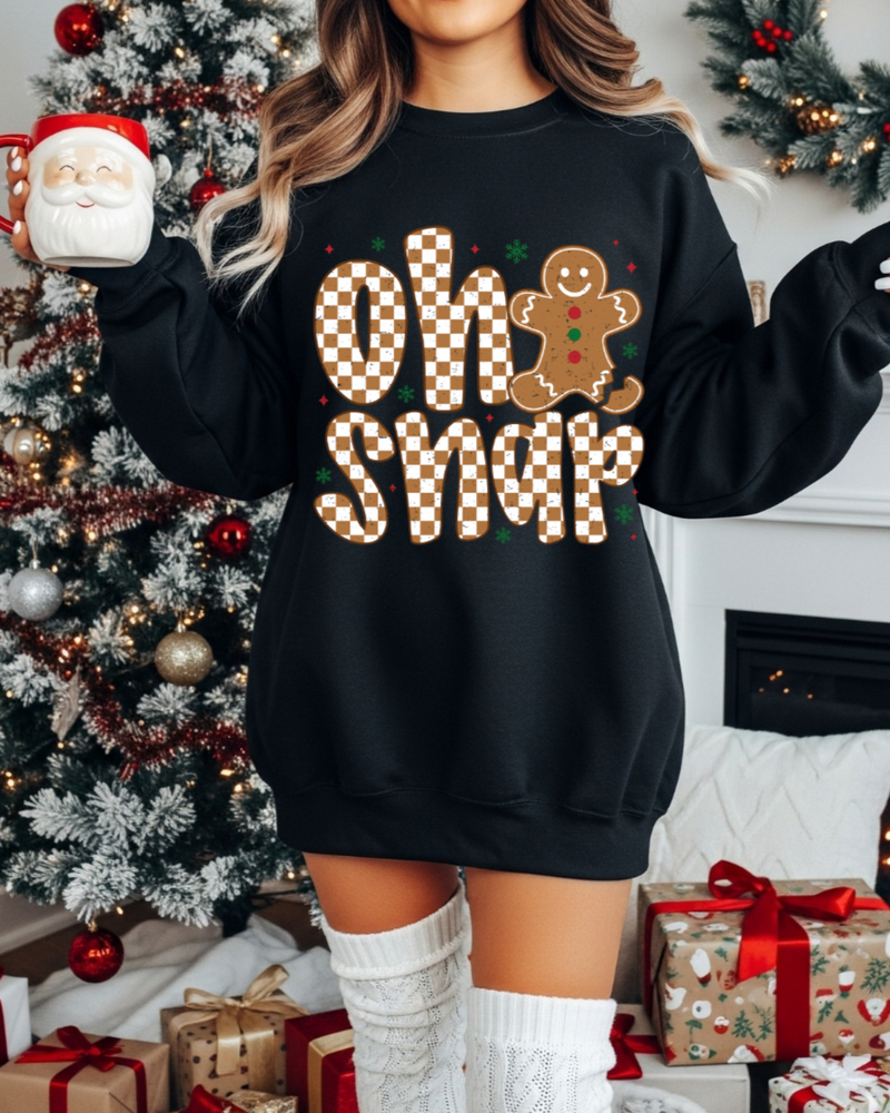 Person wearing a black sweater with 'Oh Snap' and gingerbread man design, holding a mug in a festive setting.