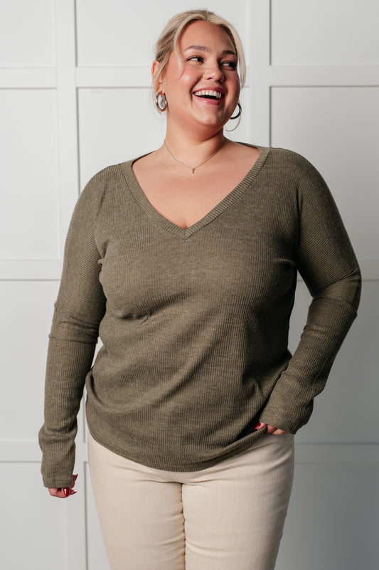 Woman wearing a green sweater and beige pants against a white tiled wall.
