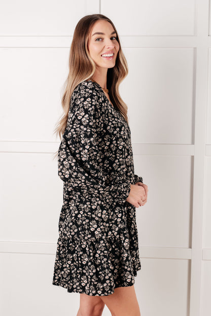 Woman wearing a black floral dress standing against a white paneled wall.