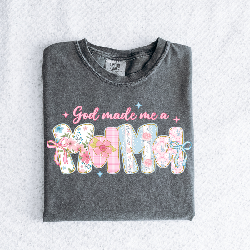 Folded dark gray t-shirt with 'God made me a MIMMA' text and floral designs on a light gray background