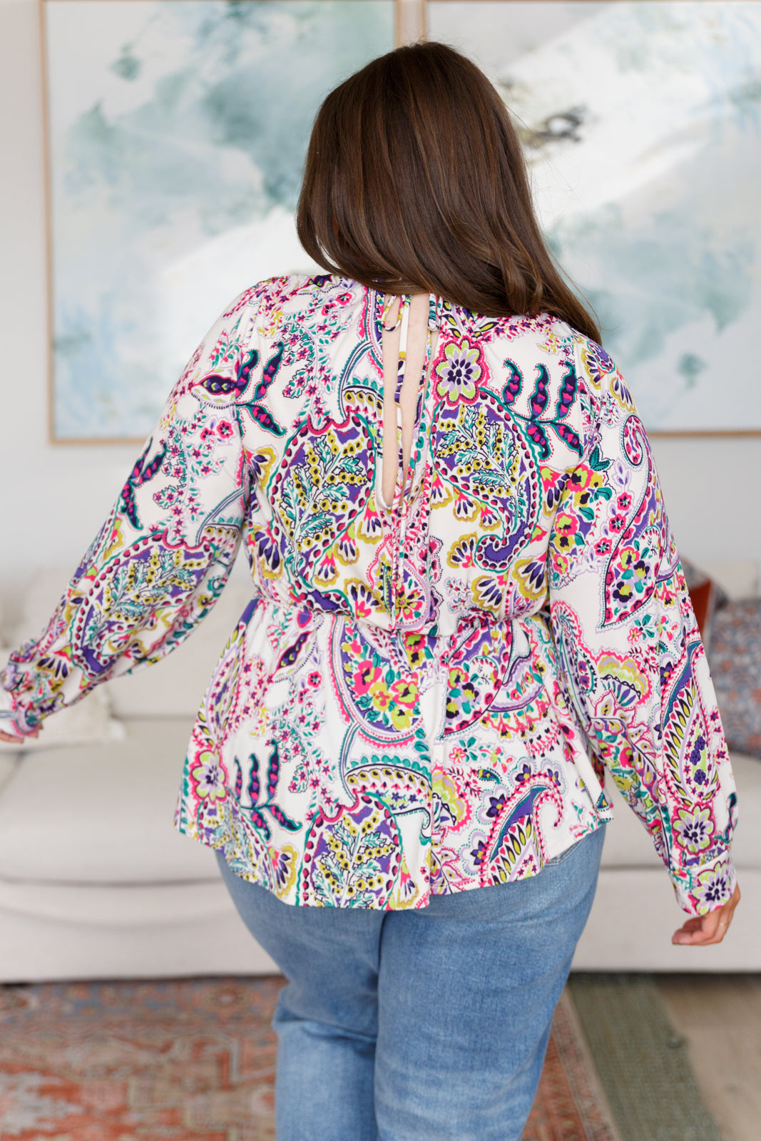 Woman wearing a colorful paisley blouse in a room with a painting on the wall.