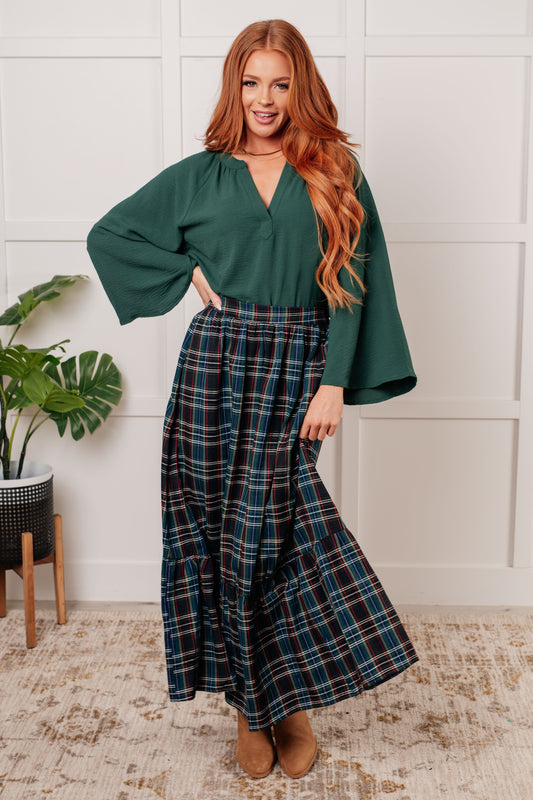 Woman wearing a green blouse and plaid pants standing in a room with a plant and rug.