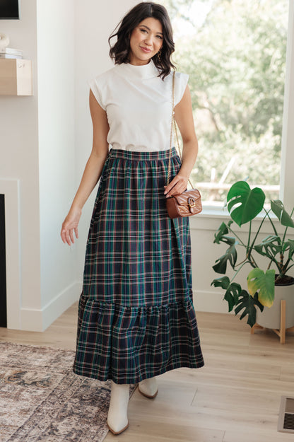 Woman wearing a white top and plaid skirt indoors with a plant in the background