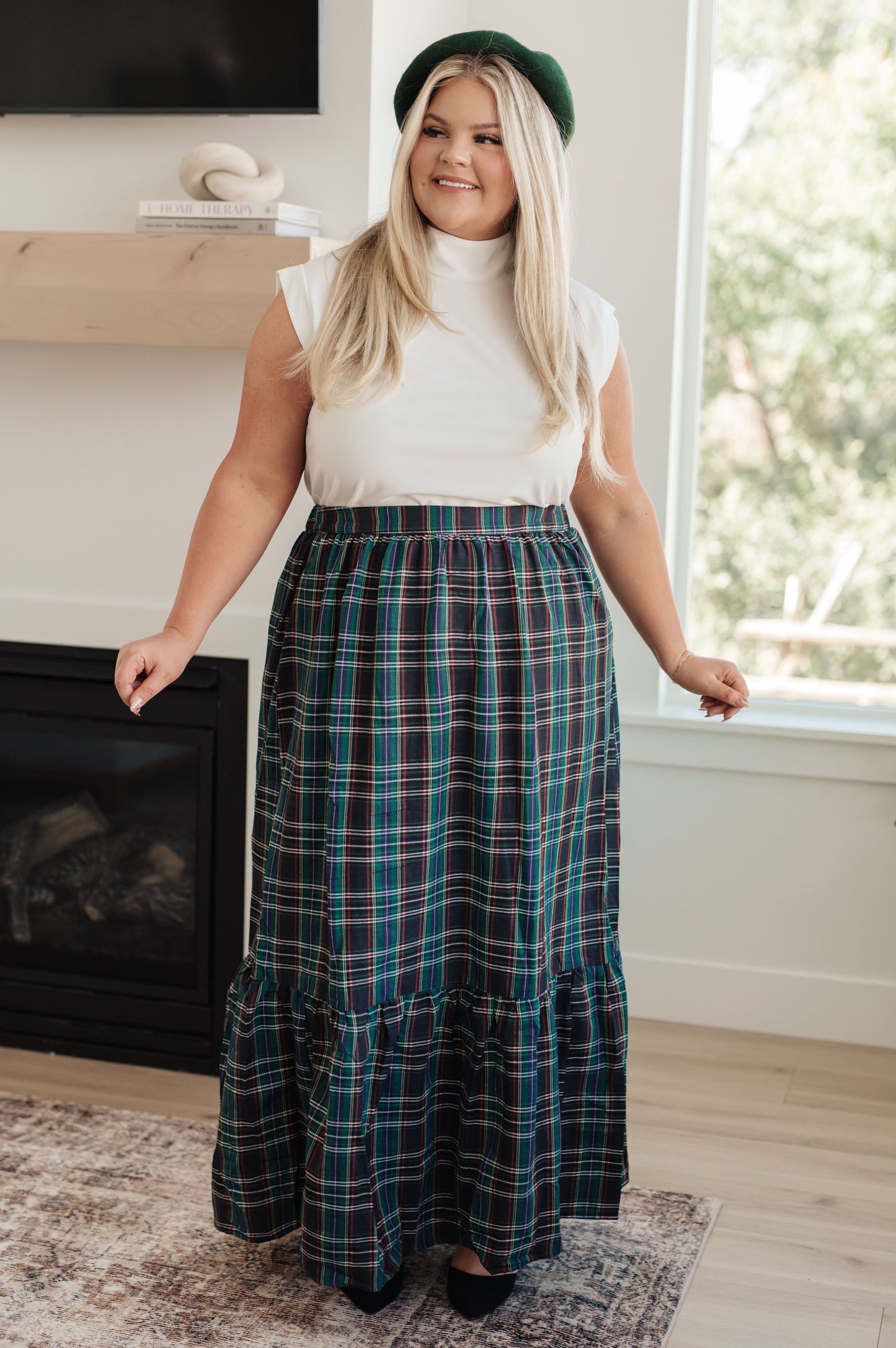 Woman wearing a white top and plaid skirt standing in a room with a fireplace and window.