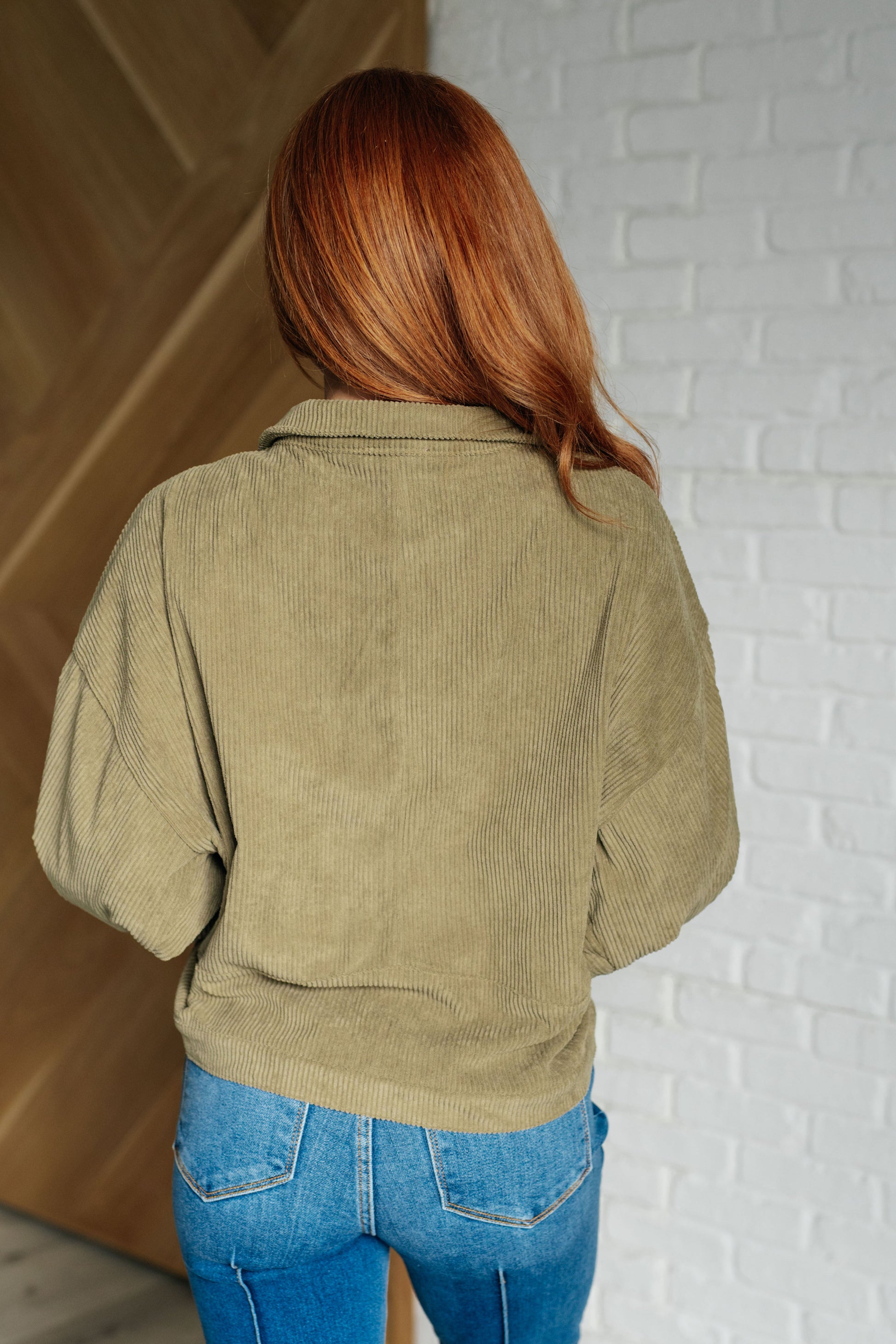 Person wearing a green corduroy jacket and blue jeans against a white brick wall.