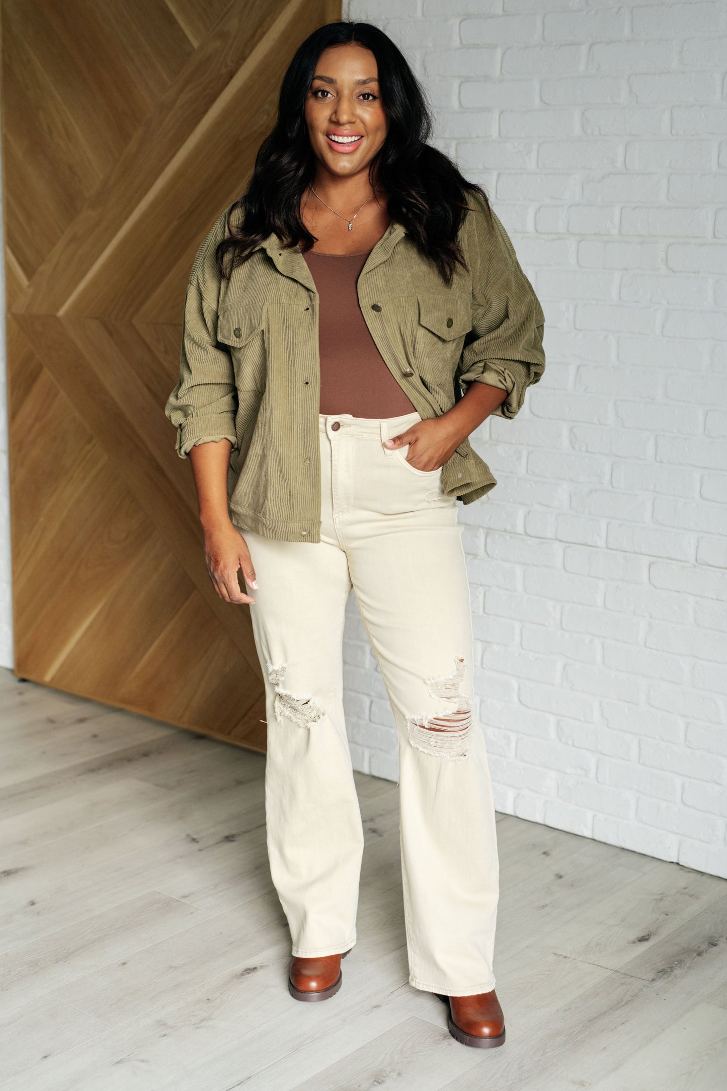 Woman wearing a green jacket, brown top, and white pants standing against a white brick wall.
