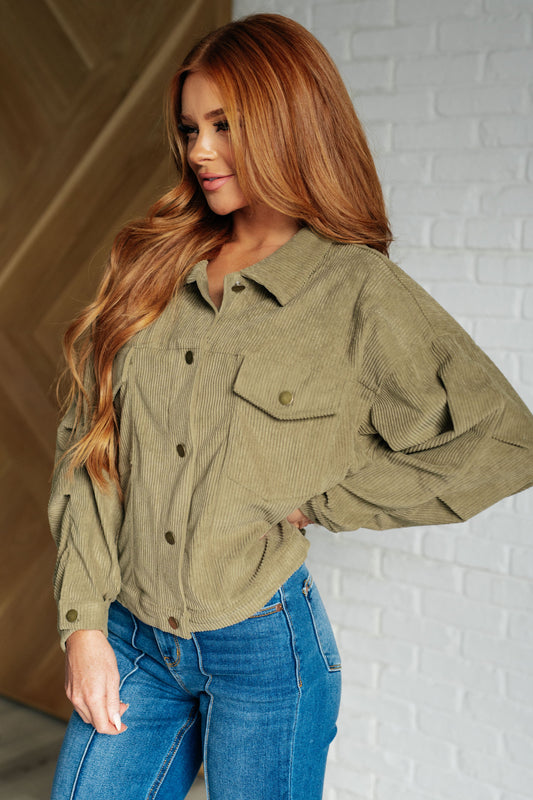 Woman wearing an olive green corduroy jacket and blue jeans against a white brick wall.