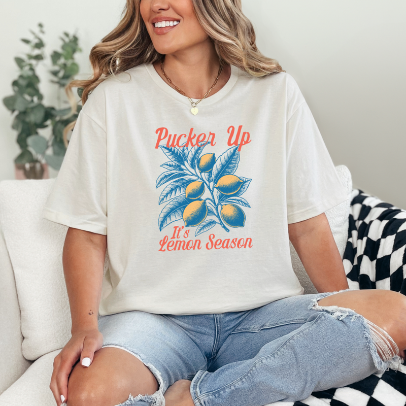 A woman sitting on a couch wearing a white graphic tee with the text 'Pucker Up It's Lemon Season' and a graphic of a lemon and palm leaves.
