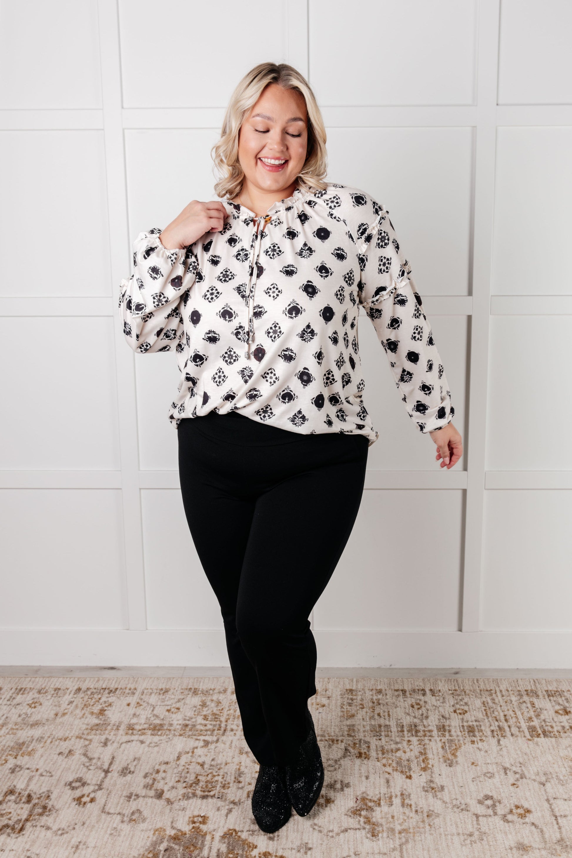 Woman wearing a patterned blouse and black pants against a white wall.
